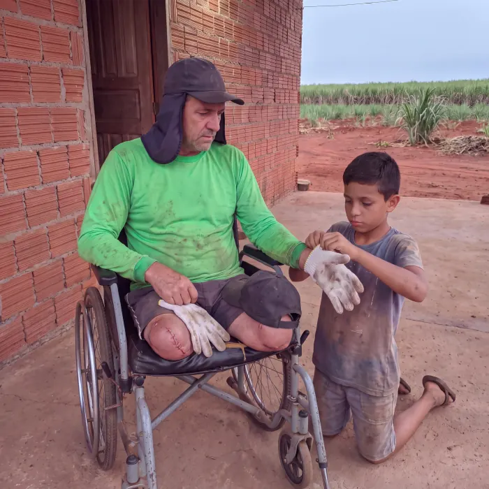 Valdir sentado na cadeira de rodas em frente a casa de tijolos, recebendo as luvas de trabalho das maos de um menino, com o canavial ao fundo