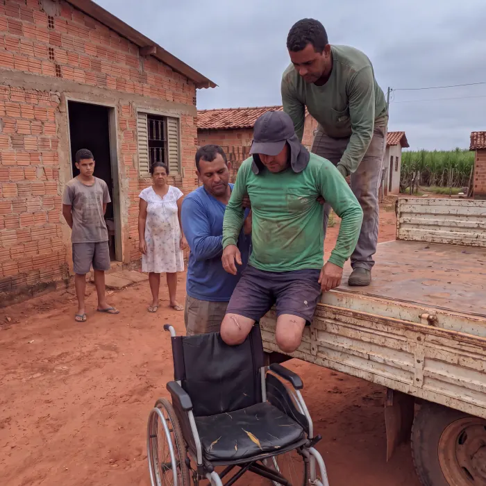 Tres homens erguem Valdir da cadeira de rodas para a carroceria do caminhao, enquanto a familia observa da porta de casa
